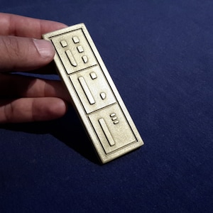 May include: A rectangular, gold-coloured metal object with three recessed sections, each containing a series of small, raised shapes. The object is held in a hand against a dark blue background.