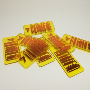 Könnte beinhalten: Eine Ansammlung gelber, rechteckiger Spielsteine mit orangefarbenem Text und Grafiken. Jeder Stein trägt die Zahl 7 und eine Reihe orangefarbener Linien. Die Steine liegen verstreut auf einer weißen Oberfläche.