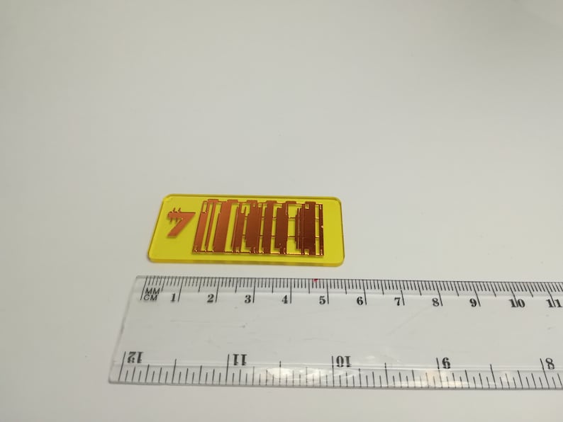 May include: A yellow rectangular badge with the number 7 and the word "MIRAGE" in orange. The badge is approximately 1.5 inches long. A clear ruler with centimeter and millimeter markings is in the foreground.