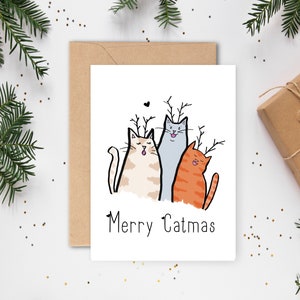 Puede incluir: Una tarjeta de felicitación blanca con tres gatos de dibujos animados con cuernos y el texto "Merry Catmas". Los gatos son de color beige, gris y naranja. La tarjeta está rodeada de ramas de hoja perenne, un sobre marrón y un regalo envuelto.