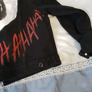 Op de afbeelding: Zwart denim jack met witte verf en rode letters die "HAHAHA" zeggen.