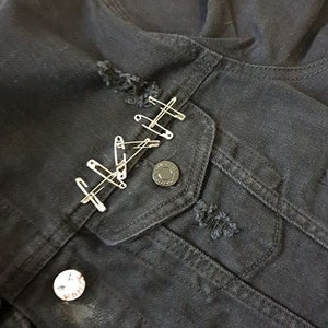 Op de afbeelding: Zwart denim jack met distressed details en veiligheidsspelden. Het jack heeft een zak met een metalen knoop en twee kleine patches.