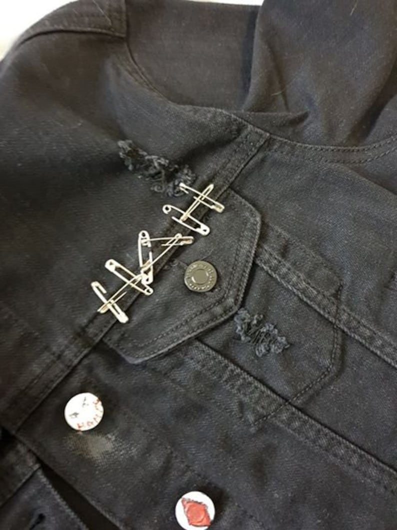 Op de afbeelding: Zwart denim jack met distressed details en veiligheidsspelden. Het jack heeft een zak met een metalen knoop en twee kleine patches.