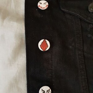 Op de afbeelding: Zwarte denimjas met drie witte emaille-pinnen. De pinnen hebben een rode ruitvorm en twee verschillende zwart-witte gezichten met de tekst "HAHAHAH" en "HAHAHA".