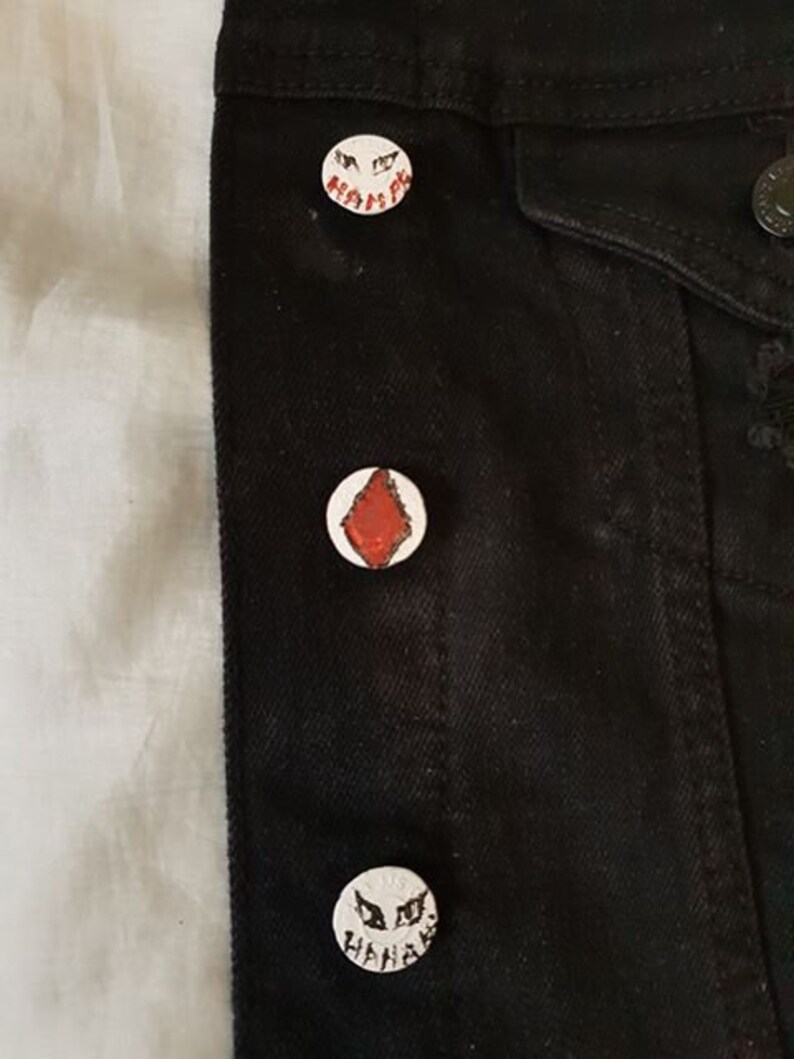 Op de afbeelding: Zwarte denimjas met drie witte emaille-pinnen. De pinnen hebben een rode ruitvorm en twee verschillende zwart-witte gezichten met de tekst "HAHAHAH" en "HAHAHA".