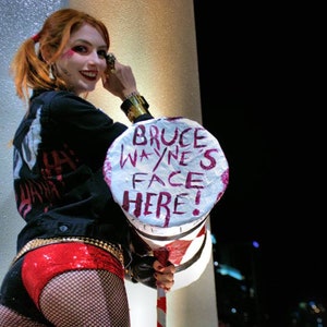 Op de afbeelding: Een vrouw verkleed als Harley Quinn houdt een wit bord vast met de woorden "Bruce Wayne's Face Here!" erop geschreven in roze. Ze draagt een zwart jack, een rode short en zwarte kousen.
