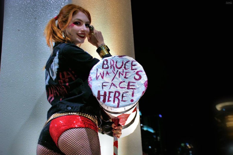 Op de afbeelding: Een vrouw verkleed als Harley Quinn houdt een wit bord vast met de woorden "Bruce Wayne's Face Here!" erop geschreven in roze. Ze draagt een zwart jack, een rode short en zwarte kousen.