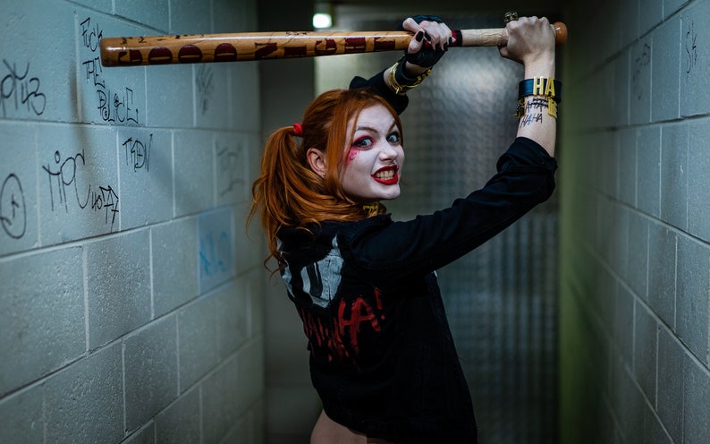 Op de afbeelding: Een vrouw verkleed als Harley Quinn, een stripfiguur, houdt een houten honkbalknuppel vast. Ze draagt een zwart jack met een witte en rode afbeelding op de rug, een rood en zwart broekje en een zwarte choker. Ze heeft rood en zwart haar, rode lippenstift en witte gezichtsverf. De achtergrond is een grijze bakstenen muur.