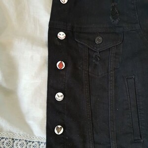 Op de afbeelding: Een zwart denim jack met verschillende witte en rode pinnen eraan bevestigd. De pinnen hebben verschillende ontwerpen, waaronder een hart, een letter J en een gezicht met ogen.