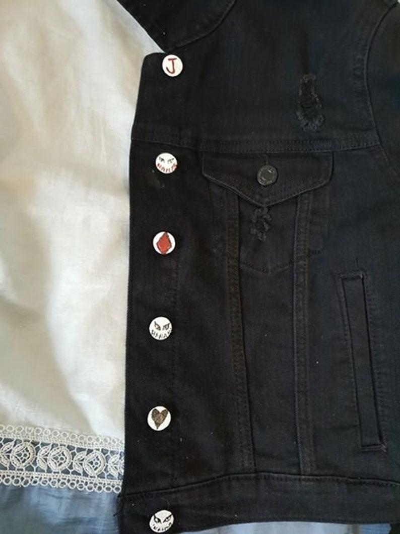 Op de afbeelding: Een zwart denim jack met verschillende witte en rode pinnen eraan bevestigd. De pinnen hebben verschillende ontwerpen, waaronder een hart, een letter J en een gezicht met ogen.