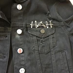 Op de afbeelding: Zwarte denimjas met verschillende pinnen en knopen, waaronder een rood-witte knop met een gezicht, een witte knop met de letter J en een witte knop met een zwart-wit gezicht. De jas heeft een distressed look met wat scheuren en gaten.