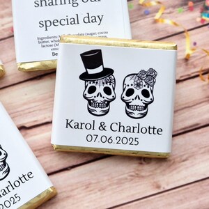 Op de afbeelding: Twee witte chocolade repen met gouden folie verpakkingen. Elke reep heeft een zwart-witte illustratie van een suiker schedel met een hoge hoed en bloemen. De tekst "Karol & Charlotte 07.06.2025" is op de reep gedrukt.