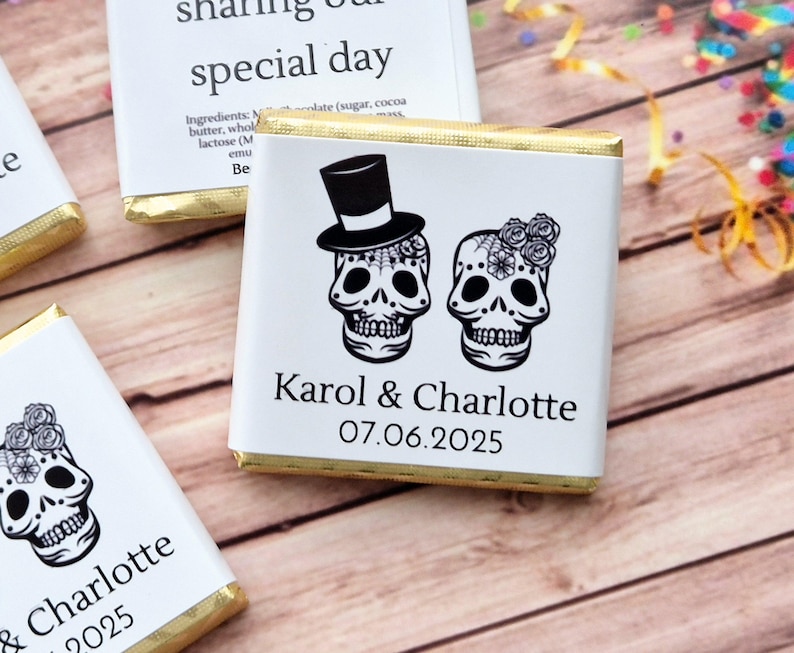 Op de afbeelding: Twee witte chocolade repen met gouden folie verpakkingen. Elke reep heeft een zwart-witte illustratie van een suiker schedel met een hoge hoed en bloemen. De tekst "Karol & Charlotte 07.06.2025" is op de reep gedrukt.