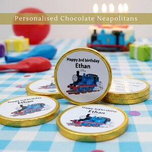 May include: Personalised chocolate Neapolitans with a gold foil edge. Each chocolate features a blue train graphic and the text "Happy 3rd birthday Ethan". Party decorations include balloons and a cake.