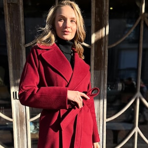 Może przedstawiać: Długi, czerwony wełniany płaszcz z zapięciem na zakładkę i paskiem do wiązania. Płaszcz jest noszony przez osobę stojącą przed drewnianymi drzwiami.