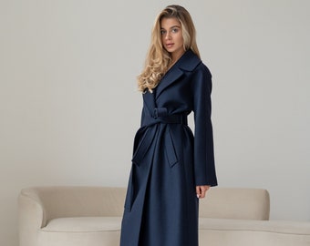 Puede incluir: Una mujer con un abrigo largo de color azul marino con cinturón. Está de pie frente a un sofá blanco y lleva pantalones blancos y mocasines negros.