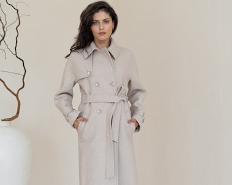 May include: A long, beige wool coat with a double-breasted closure, a belt, and pockets. The coat is worn by a person with long dark hair and is standing in a white room.
