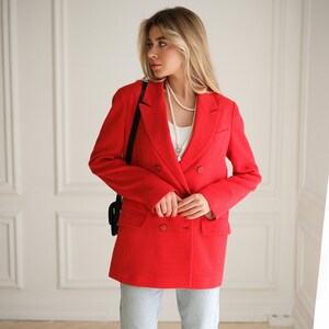 May include: A woman wearing a red double-breasted blazer with a pearl necklace and light blue jeans.