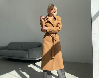 May include: A long camel-coloured coat with a belt and a double-breasted closure. The coat is being worn by a person with blonde hair and is standing in a white room.