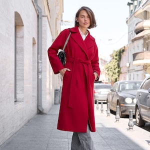 Puede incluir: Un abrigo largo de color rojo vibrante en mezcla de lana con solapa ancha, mangas largas y un cinturón de tela a juego en la cintura. El abrigo se lleva sobre una camisa blanca de cuello y pantalones grises de pierna ancha, con un bolso negro.