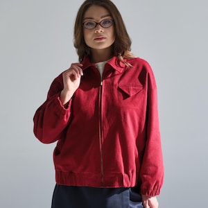 May include: A woman wearing a red zip-up jacket with a dark blue skirt and glasses.