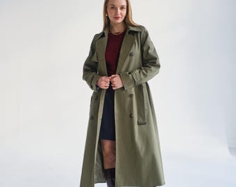 May include: A woman wearing a long, olive green trench coat with a double-breasted closure and a belt. She is also wearing a burgundy top and dark blue skirt. The woman is wearing brown heeled boots.