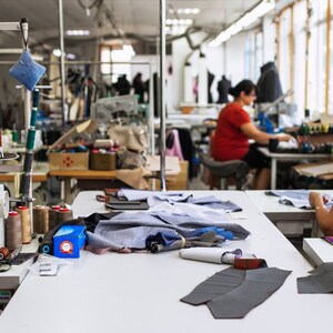May include: A sewing workshop with two people working on sewing machines. One person is using a white sewing machine with spools of thread and fabric scraps on the table. The other person is using a black sewing machine with a spool of thread and fabric scraps on the table.