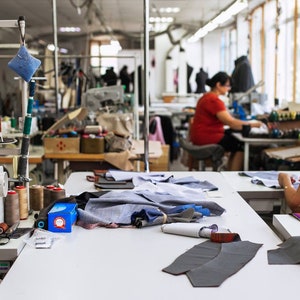 May include: A sewing workshop with two people working on sewing machines. One person is using a white sewing machine with spools of thread and fabric scraps on the table. The other person is using a black sewing machine with a spool of thread and fabric scraps on the table.