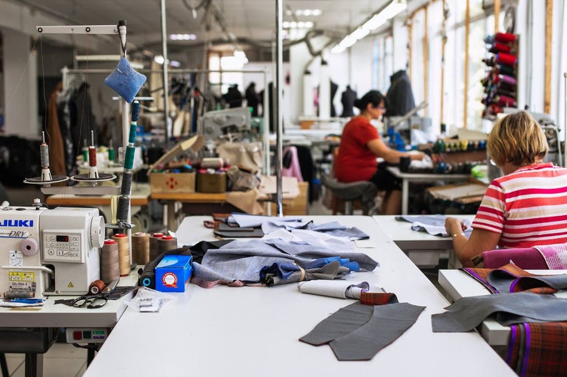 May include: A sewing workshop with two people working on sewing machines. One person is using a white sewing machine with spools of thread and fabric scraps on the table. The other person is using a black sewing machine with a spool of thread and fabric scraps on the table.
