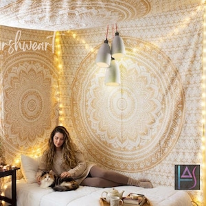 May include: A bedroom scene featuring a white bed with a white comforter, a decorative gold and white mandala tapestry, and string lights. The text "Harshwear" is visible. A person is sitting on the bed with a cat.