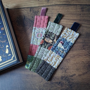 May include: Three fabric bookmarks with velvet ribbon tabs on a wooden surface next to a book. The bookmarks display floral and geometric patterns in pink, green, black, and gold. The book has a dark blue cover with gold detailing.