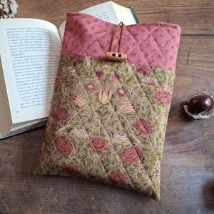 May include: A quilted book sleeve featuring a floral design in pink, green, and beige. The top section is a solid pink with a diamond pattern. It has a wooden toggle closure.