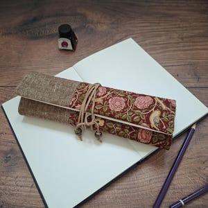 May include: A fabric pencil roll featuring a brown linen section and a floral pattern with birds in shades of red, green, and cream. The roll is secured with a thin leather cord tied around the centre, finished with two small wooden beads at the ends.