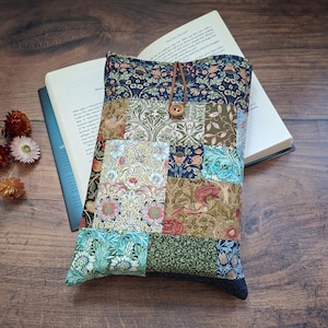 May include: A rectangular fabric sleeve featuring a patchwork design of various floral and botanical patterns in shades of navy blue, green, pink, and tan. The sleeve includes a brown leather cord closure secured by a small wooden button.
