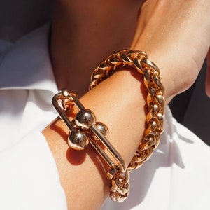 May include: Gold-toned bracelets. One bracelet features a thick, braided chain design. The other bracelet has a unique design with a large clasp and three spherical accents. The jewelry is worn on a wrist, against a white shirt.