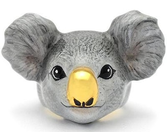May include: A gray ceramic koala ring with a gold nose and black eyes. The ring is a realistic depiction of a koala.