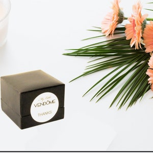 May include: A small black gift box with a white circular sticker that reads "My Little VENDOME THANKS". The box is on a white surface with a bunch of pink gerbera daisies and green palm leaves.