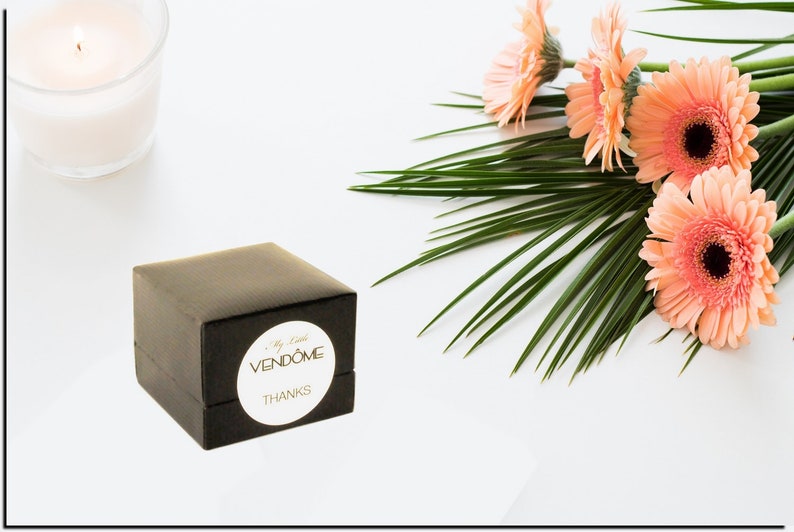 May include: A small black gift box with a white circular sticker that reads "My Little VENDOME THANKS". The box is on a white surface with a bunch of pink gerbera daisies and green palm leaves.