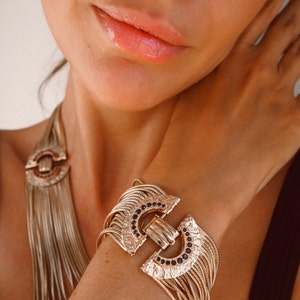 Peut inclure: Un collier et un bracelet en chaîne dorée avec un fermoir décoratif présentant un motif géométrique avec des pierres précieuses noires. Les bijoux sont superposés et ont une esthétique vintage.