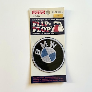 Op de afbeelding: Een zwart-witte geborduurde strijkpatch met het BMW-logo. De patch is verpakt op een witte kaart met de tekst "Flip-Flop" en "Gold-Zack" erop gedrukt.