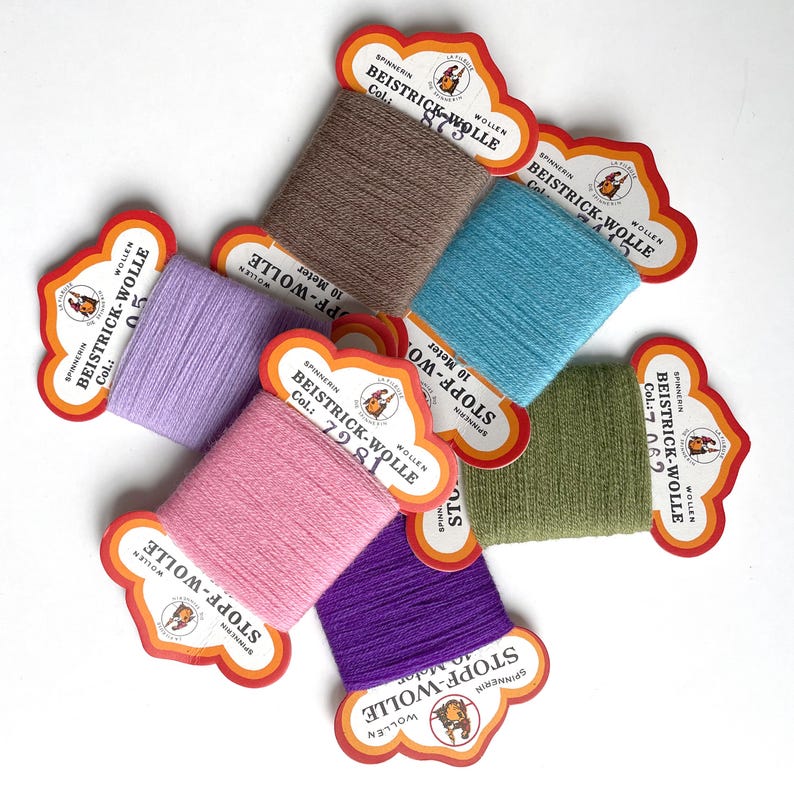May include: Six spools of yarn in various colours, including purple, pink, light green, light blue, brown, and lavender. Each spool has a label with the text "BEISTRICK-WOLLE" and "STOPF-WOLLE" in German. The labels also include the colour number and the text "SPINNERIN WOLLEN".