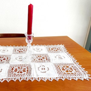 May include: A red candle sits in a clear, ornate glass candlestick on a white, square-patterned lace runner. The runner is placed on a wooden table. The candle is tall and slender, and the lace has a scalloped edge.