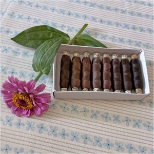 May include: A white cardboard box containing eight spools of dark brown sewing thread, each with a paper label on the top. A single pink zinnia flower with a yellow center and two green leaves is positioned next to the box of thread.