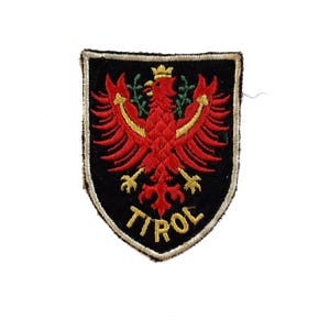 Könnte beinhalten: Gestickter Aufnäher mit einem roten Adler mit goldener Krone und Olivenzweigen auf einem schwarzen Schild. Das Wort "TIROL" ist in Gold gestickt. Der Aufnäher hat einen schwarzen Rand.