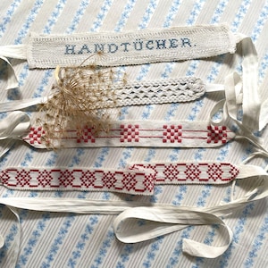 May include: Four vintage textile bands with white ribbons. One band has the word "HANDTÜCHER" embroidered in blue. Two bands feature red and white geometric patterns. The fourth band is a white open weave.