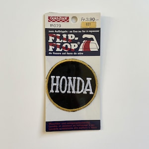 May include: A black and white iron-on patch with the word "HONDA" in white lettering on a black background. The patch is surrounded by a gold border and is packaged on a white card with the words "Flip-Flop" and "Gold-Zack" printed on it.