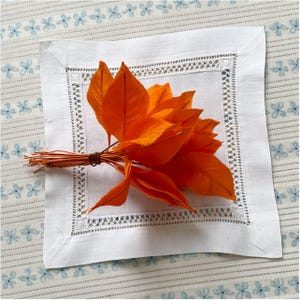 Könnte beinhalten: Ein Bündel leuchtend orangefarbener Blätter, mit Bindfaden zusammengebunden, liegt auf einer weißen Leinen-Serviette mit zarter Spitzenbordüre. Die Serviette befindet sich auf einer hellblauen, floral gemusterten Oberfläche.