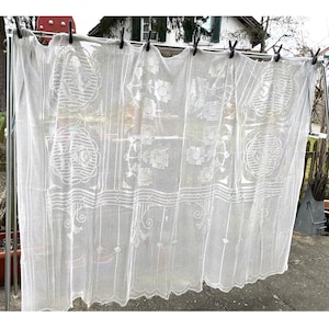 May include: White lace curtain with floral and geometric patterns, held by black clothespins on a metal rod. The curtain has a scalloped bottom edge and is semi-transparent, allowing light to pass through. The design includes large floral motifs and horizontal lines.