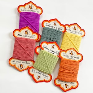 Op de afbeelding: Assortiment van kleurrijke garenklossen met etiketten in het Frans. De etiketten luiden "Laine à Renforcer", "Laine à Repriser", "Beistrick-Wolle" en "Laine & Renforcer".