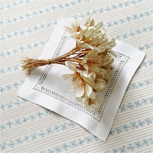 May include: A white linen napkin with a decorative cutwork border, topped with a bundle of cream-colored artificial flowers tied with twine. The napkin rests on a patterned tablecloth with blue floral stripes.